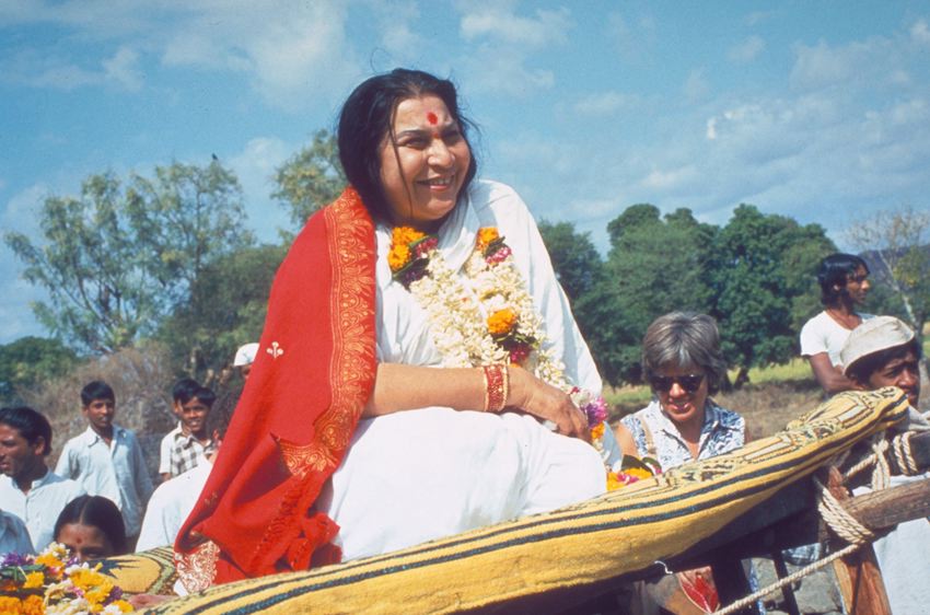 Shri Mataji Nirmala Devi sitting on a cart during a procession in India, garlands, white sari, red shawl